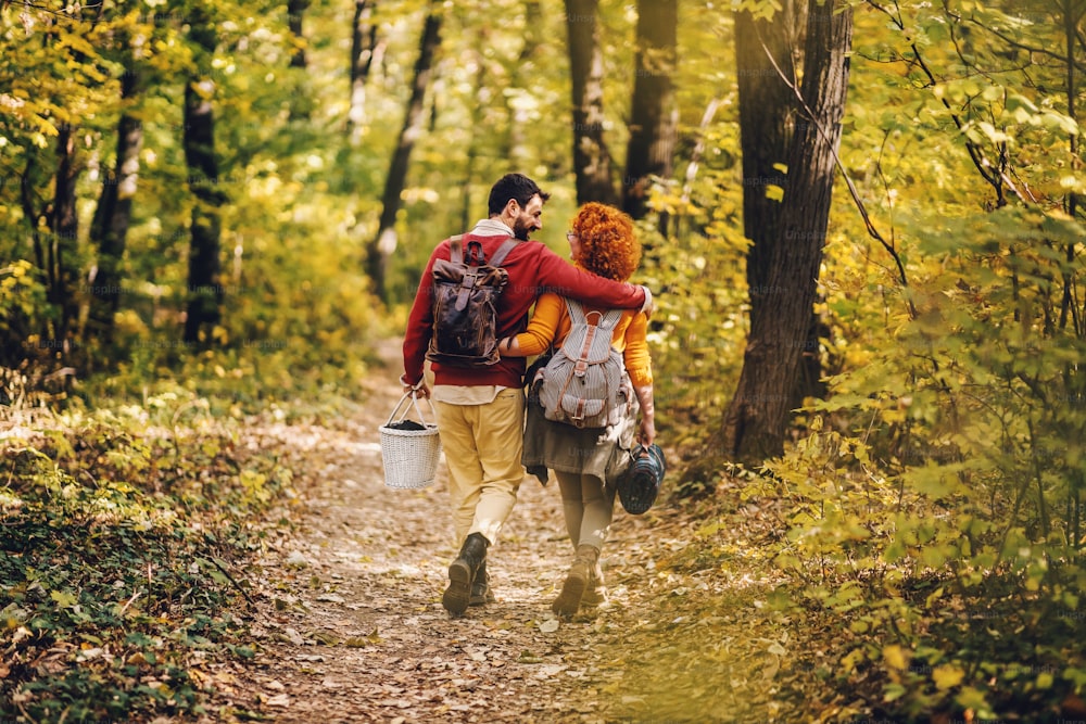 Rear view of young happy couple in love hugging and walking in nature on a beautiful autumn day. Couple holding picnic equipment.