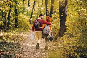 Rear view of young happy couple in love hugging and walking in nature on a beautiful autumn day. Couple holding picnic equipment.