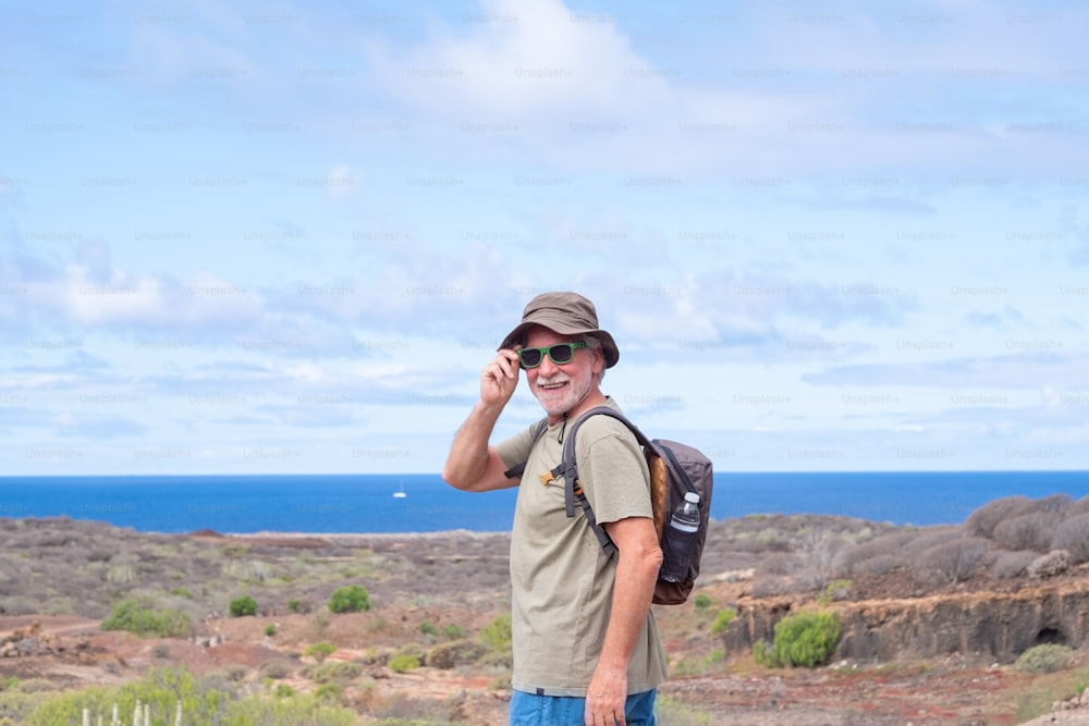 Happy active senior man with backpack and hat while hiking outdoors looking at camera smiling. Elderly man enjoying retirement and healthy lifestyle. Blue sea and sky in the background