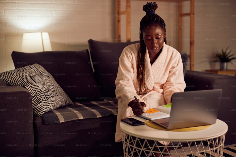 Mulher afro-americana sorridente usando laptop e escrevendo notas enquanto trabalha até tarde em casa.