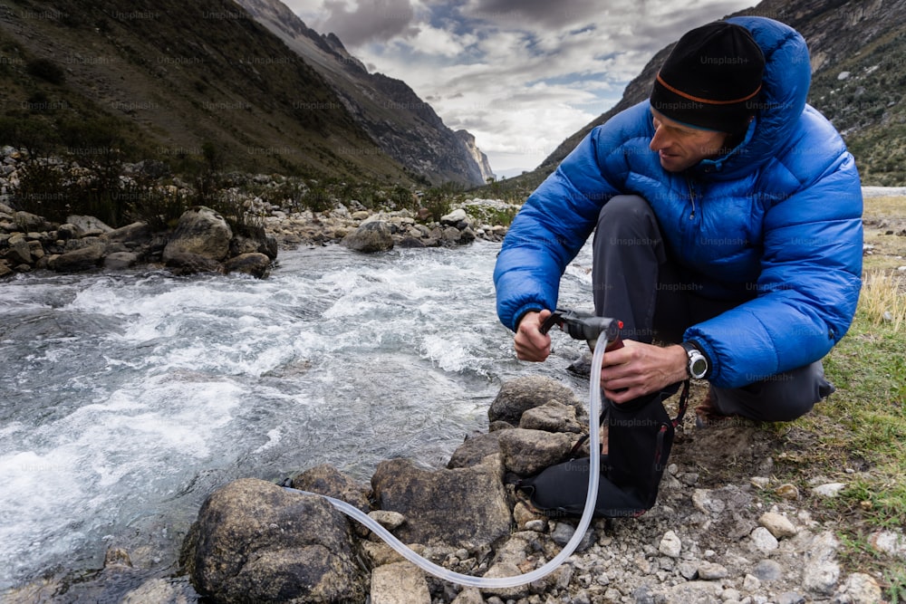 青いダウンジャケットを着た男性バックパッカーが、ペルーのアンデス山脈にあるコルディレラ・ブランカの野生の渓流から飲料水をろ過する