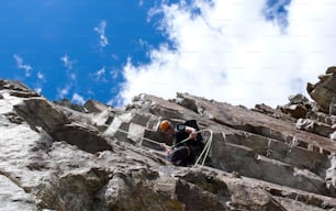 A male mountain guide rappelling off a steep rock face under a beautiful blue sky with white clouds