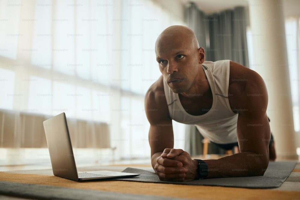 Homem atlético afro-americano que exercita resistência em pose de prancha enquanto treina esportes em casa.
