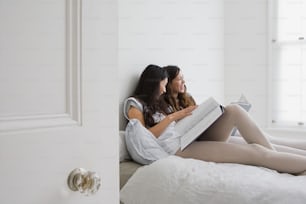 two women are sitting on a bed reading a book