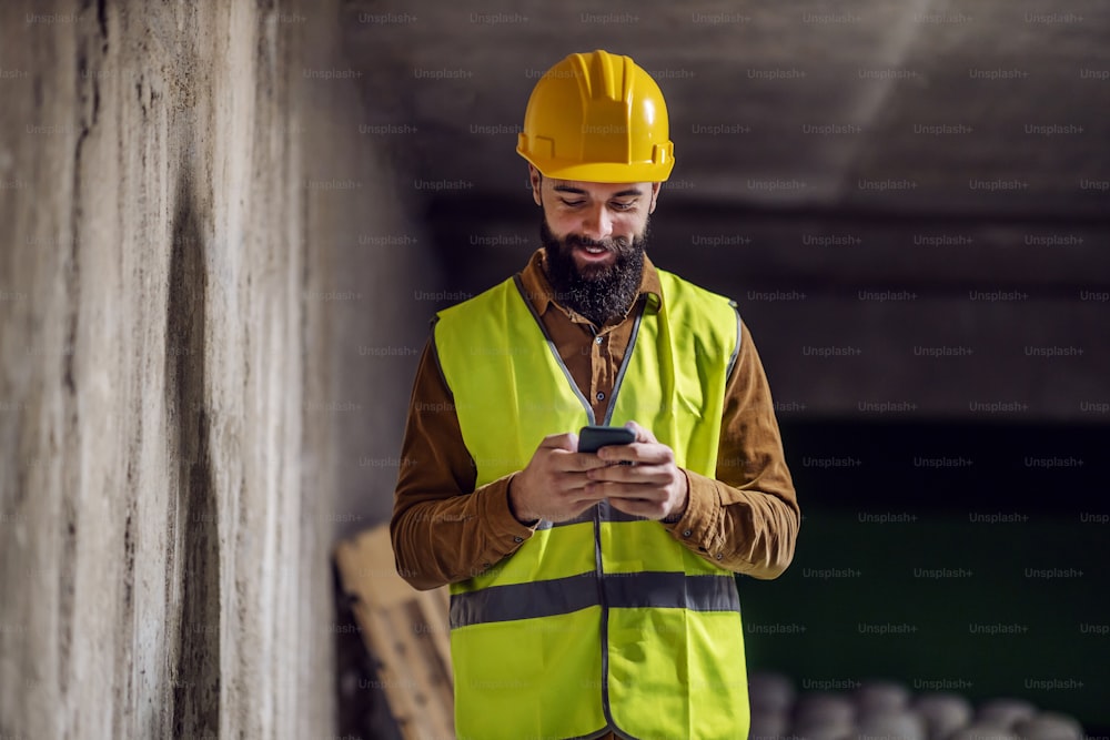 Jovem atraente trabalhador barbudo sorridente em colete com capacete na cabeça em pé dentro do prédio em processo de construção, digitando uma mensagem no telefone inteligente e fazendo uma pausa no trabalho.