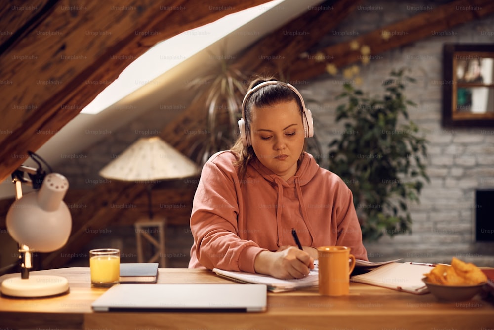 Female university student wearing headphones and writing in notebook while learning at home.