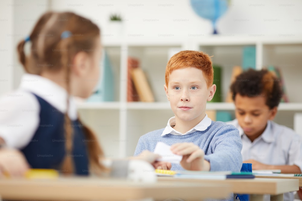 Red haired schoolboy sitting at desk and giving a note to the schoolgirl during a lesson at school