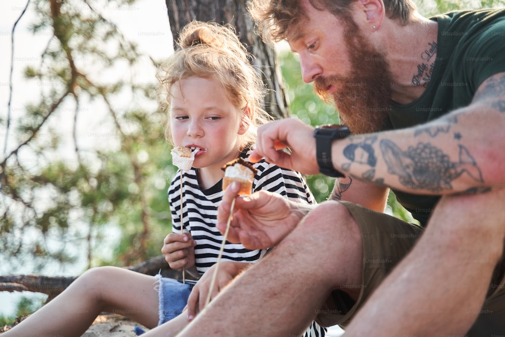 Tasty dinner. Father and daughter camping together. They eating tasty fried marshmallow and chatting with each other. Family concept