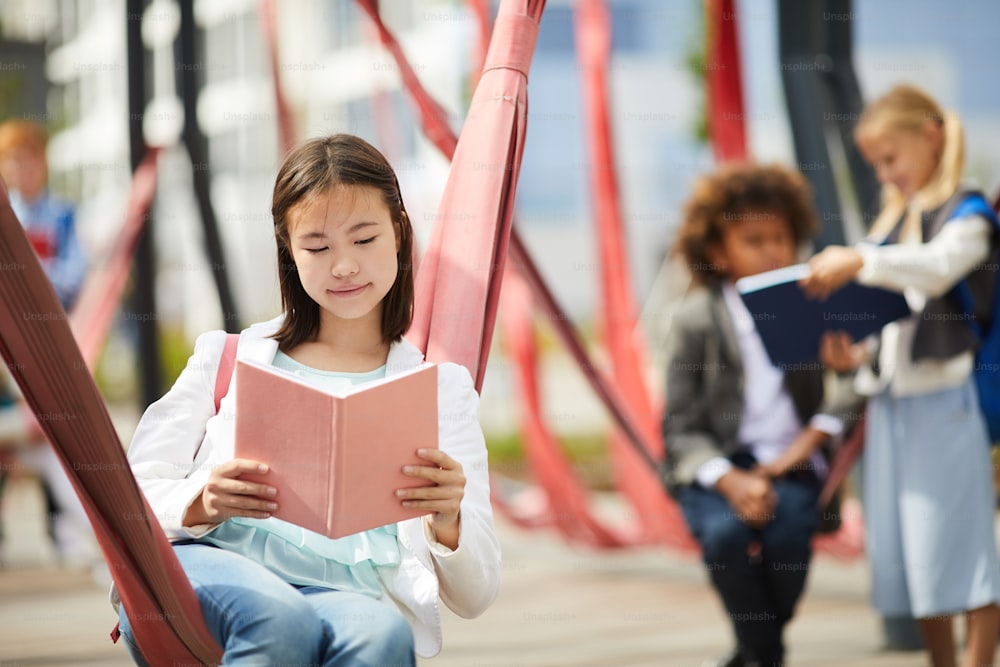 Asian schoolgirl reading a book while sitting at schoolyard outdoors with her classmates in the background
