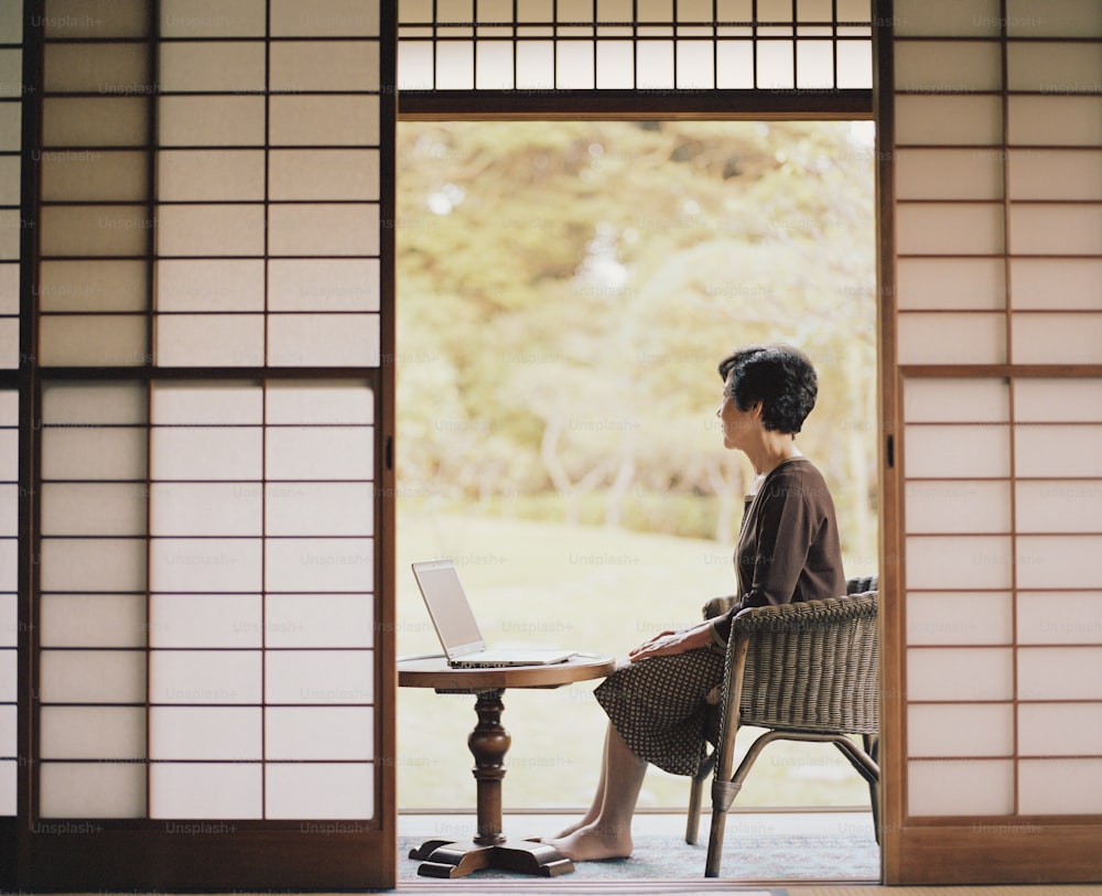 Mujer mayor sentada al lado de la puerta abierta, usando computadora portátil, vista lateral