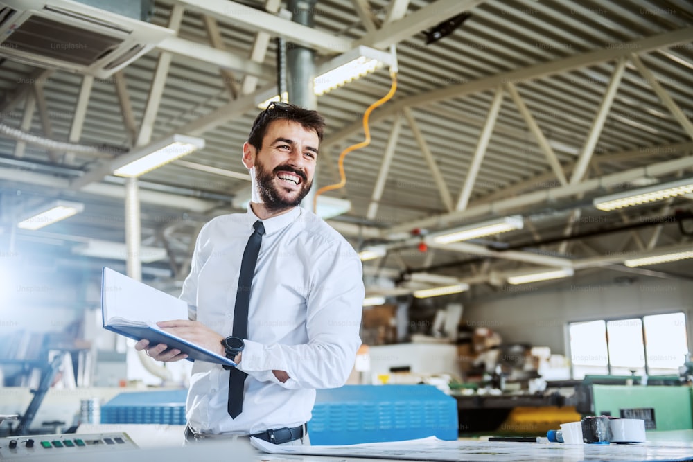 Handsome smiling caucasian bearded graphic engineer holding notebook while standing in printing shop. In background are printing machines.