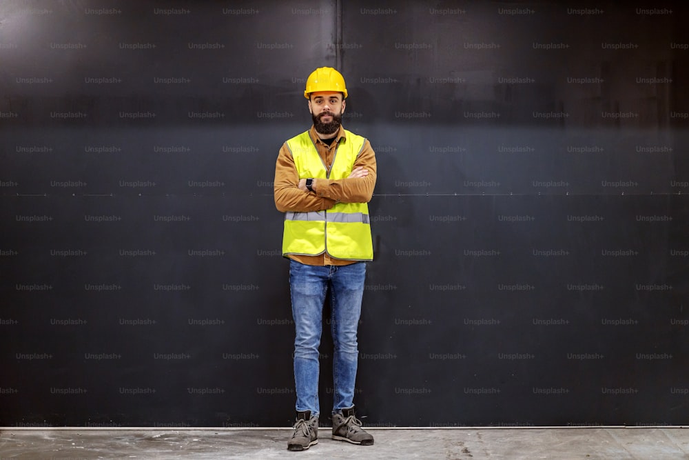 Young smiling attractive bearded worker in vest with helmet on head standing in front of black door with arms crossed and looking at camera.