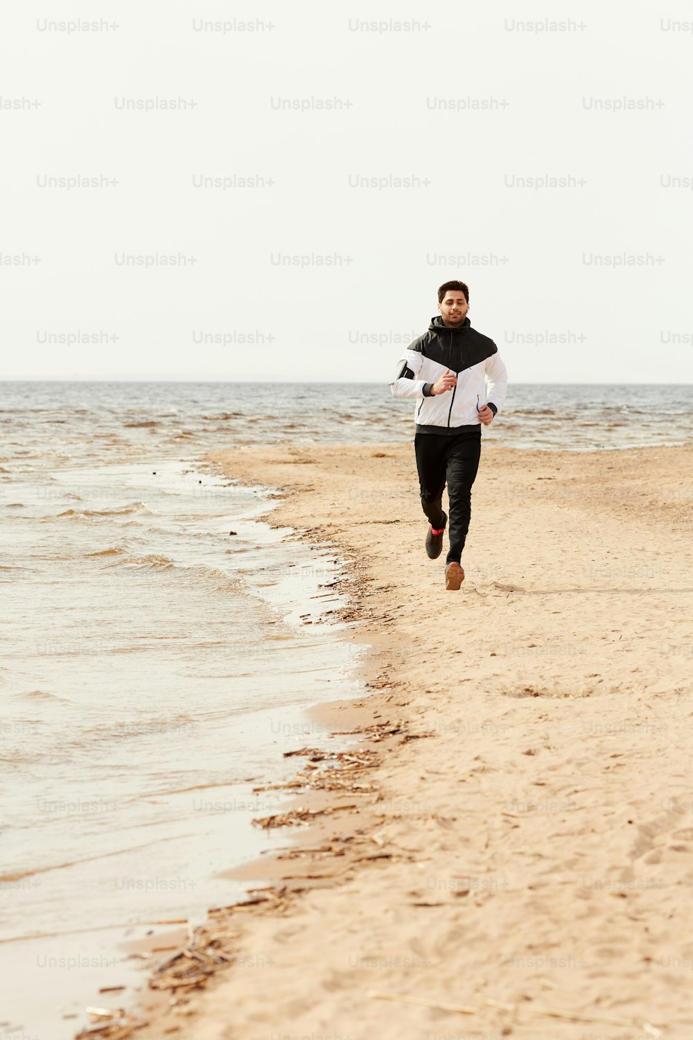 Young sportsman in activewear running down sandy beach along coastline on cool summer day or morning