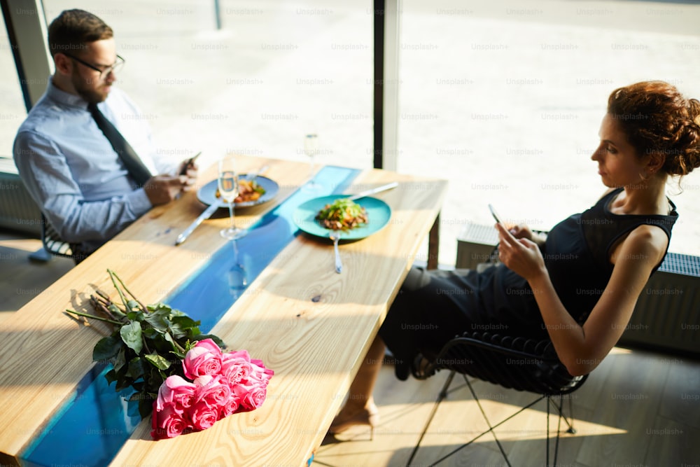 Jeune homme et femme contemporains dans des vêtements élégants assis près de la table servie dans le restaurant et utilisant des gadgets