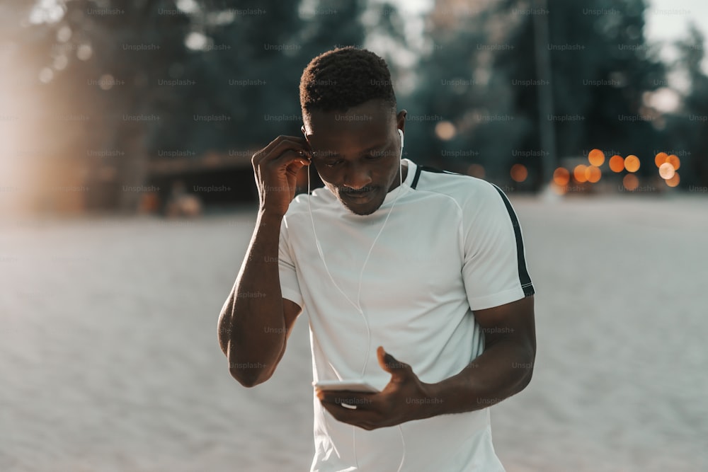 Fit african man in white t-shirt putting earphones in ears and holding smart phone.