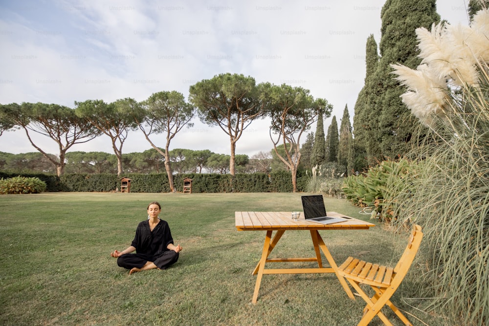 Young caucasian girl meditating and practicing yoga on green lawn of mediterranean yard. Concept of weekend, rest and vacation. Idea of healthy lifestyle. Wooden chair and table with laptop