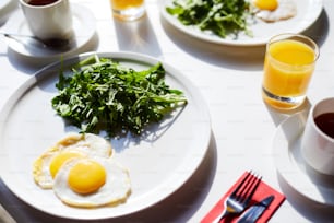 Close-up of fried eggs with greens on plate, cup of tea, silverware and glass of juice for breakfast