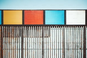 Vista frontal de uma parede do edifício coberta por muitas listras verticais de madeira e áreas de retângulo de metal no topo preenchidas por cores sólidas vívidas, com uma faixa de céu claro acima