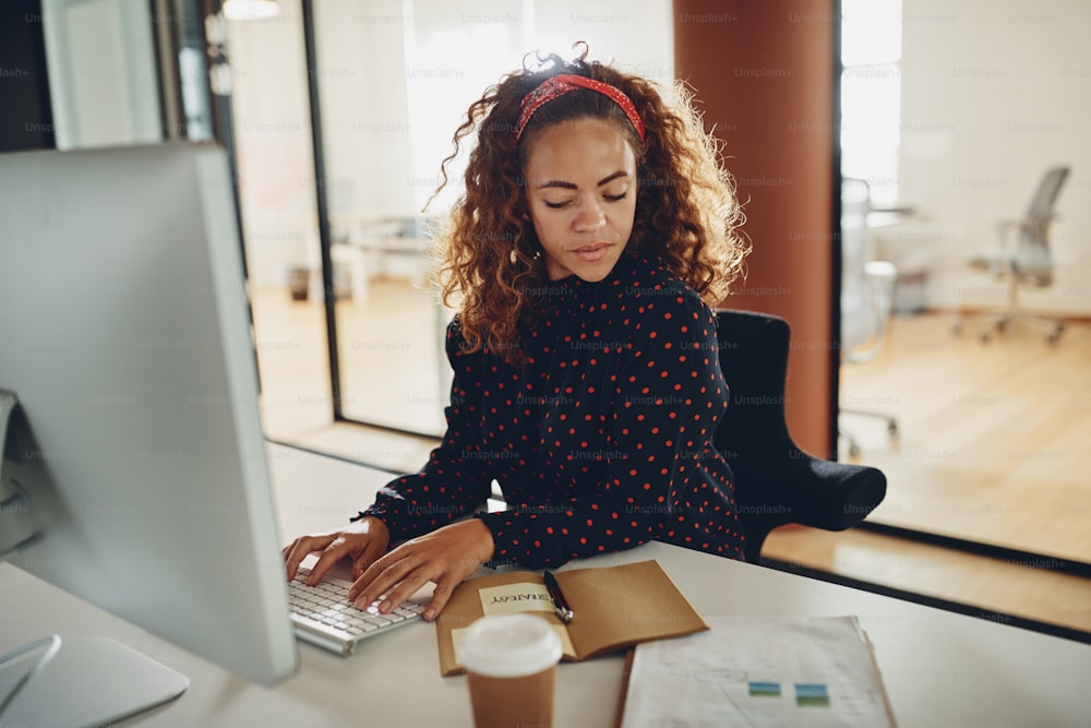 Junge Geschäftsfrau, die an ihrem Computer arbeitet und Papierkram liest, während sie an einem Schreibtisch in einem Büro sitzt