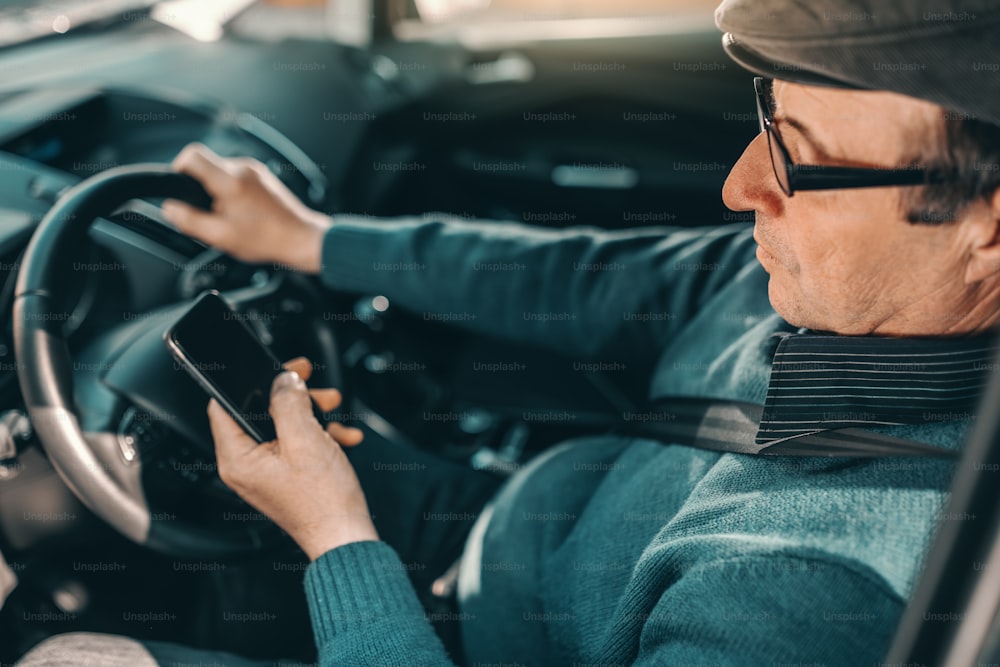 Kaukasischer Senior mit Hut auf dem Kopf und Brille fährt Auto und benutzt Smartphone.
