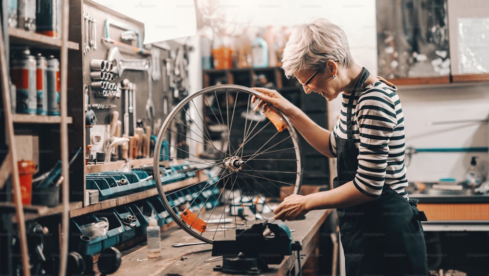 Cute Caucasian female worker holding and repairing bicycle wheel while standing in bicycle workshop.