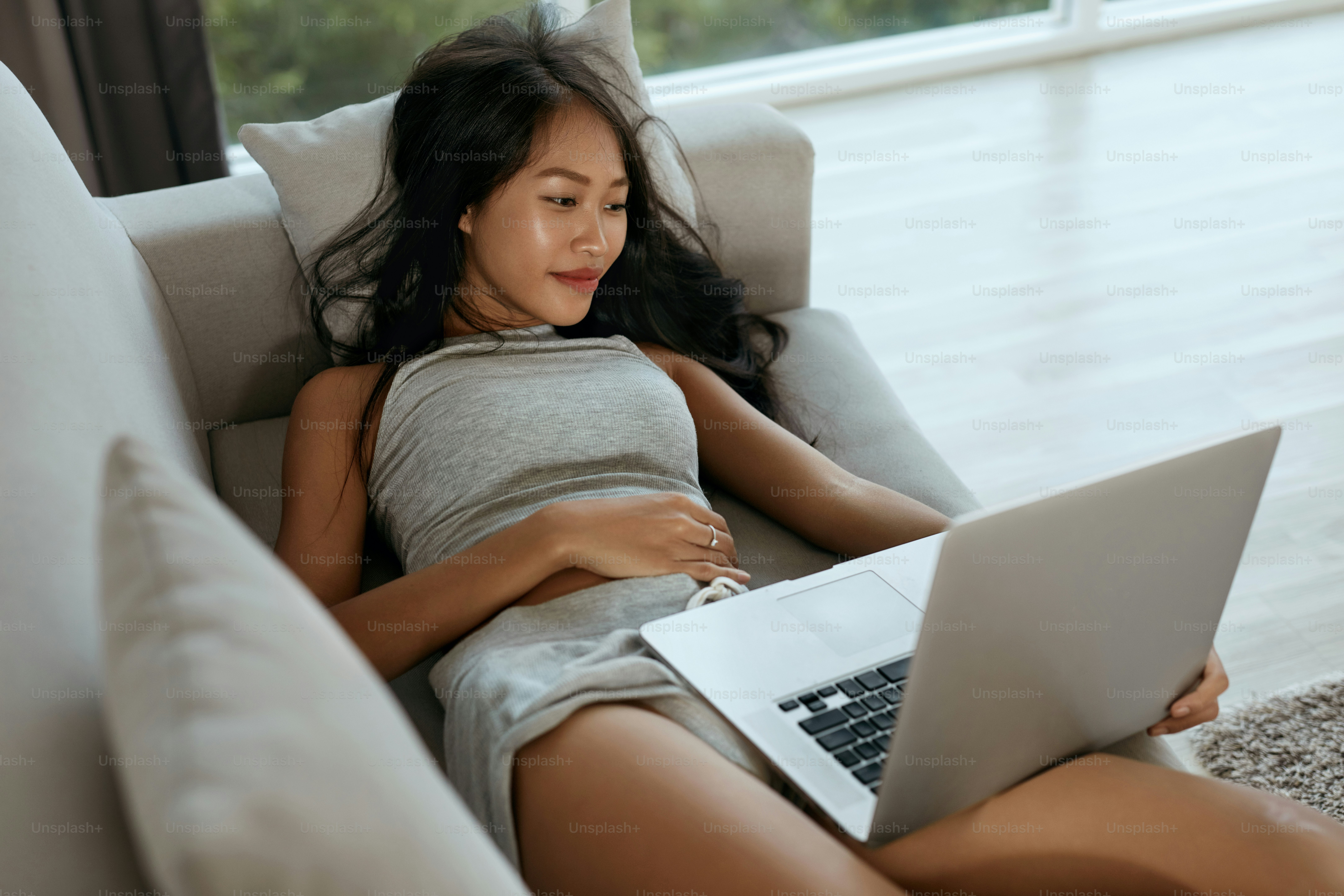 Woman with computer on couch at home in morning. Beautiful smiling asian girl using laptop relaxing on sofa in light living room