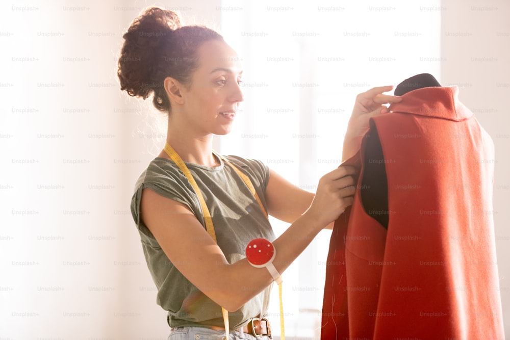 Junge Frau in Freizeitkleidung steht mit rotem Mantel vor einem Schnuller und probiert ihn bei Näharbeiten an