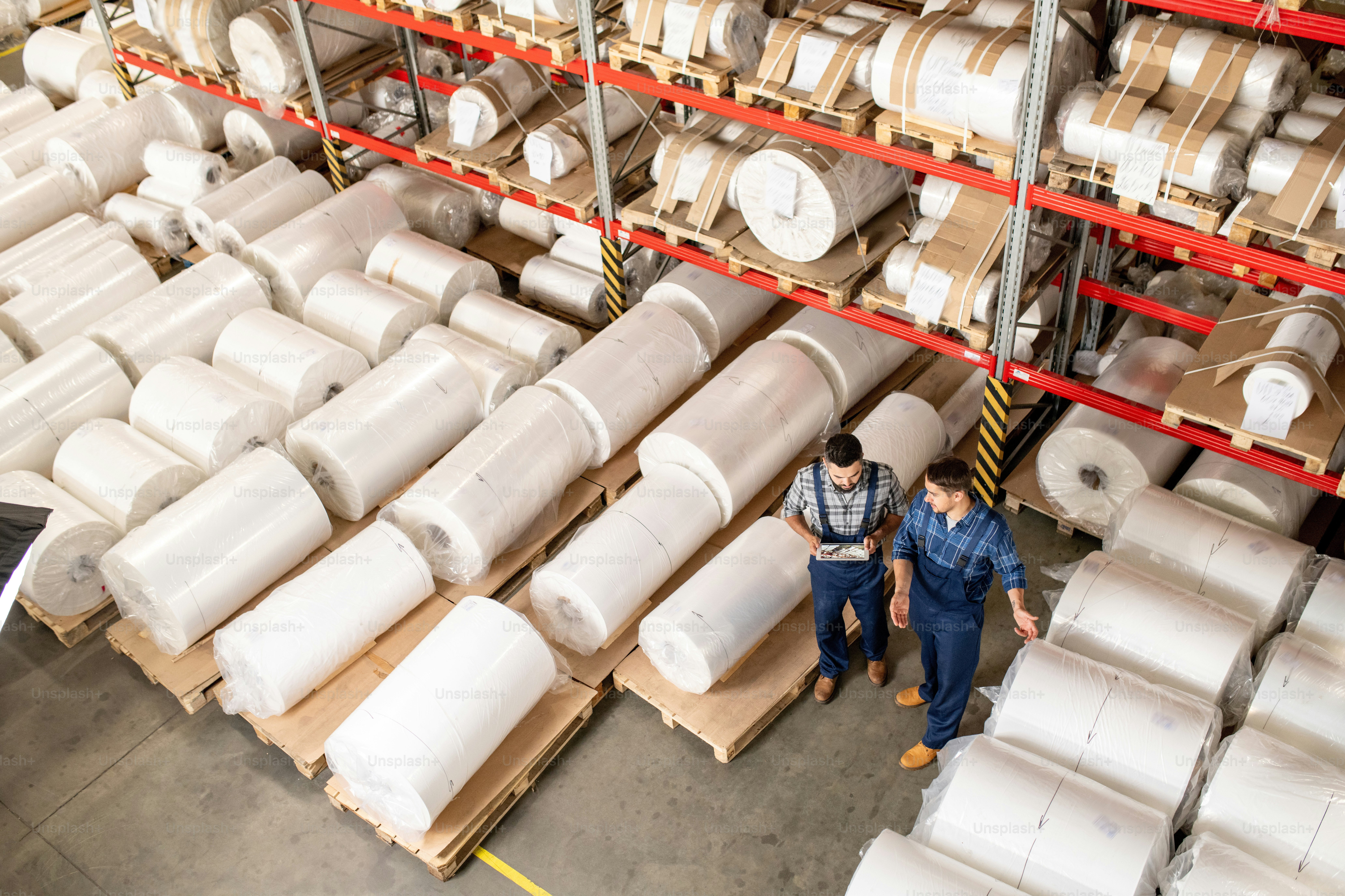 Two professional engineers in workwear discussing points of quality control in warehouse of large processing plant