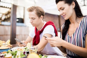 Side view portrait of two modern young people texting at table in cafe, focus on woman in front using smartphone, copy space