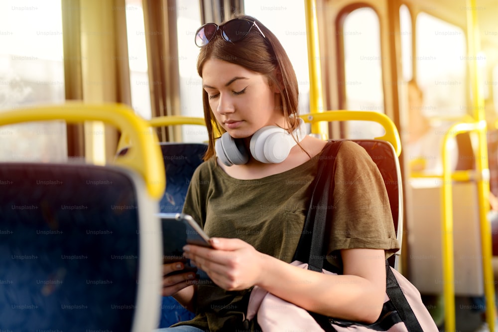 Serious young student girl sitting in a bus and looking at her telephone with serious look. Holding headphones around her neck.