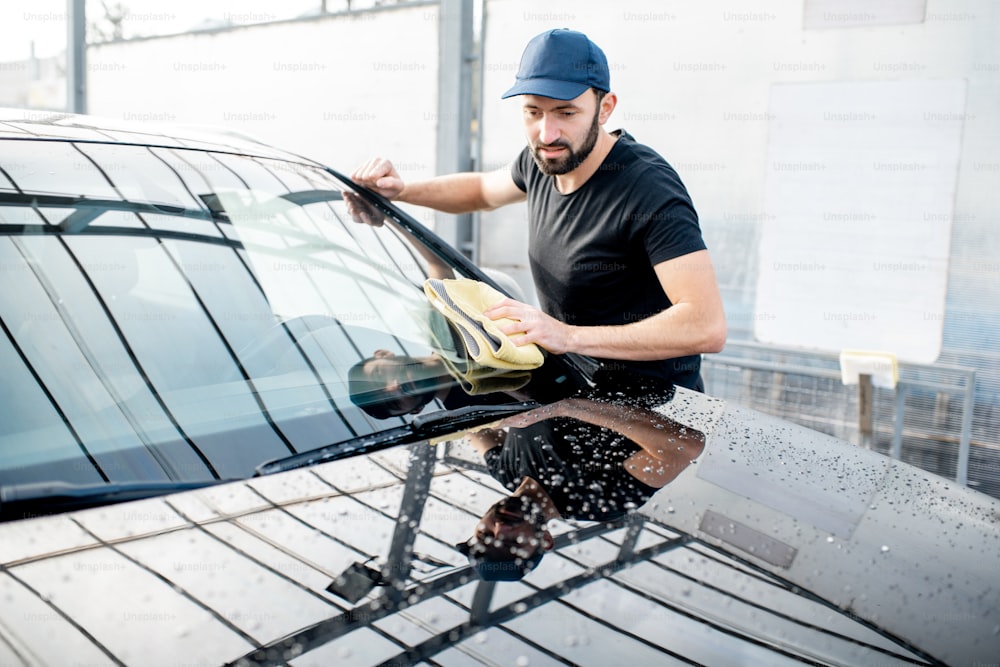 Professional washer in t-shirt and cap wiping windshield with yellow microfiber at the open air car wash