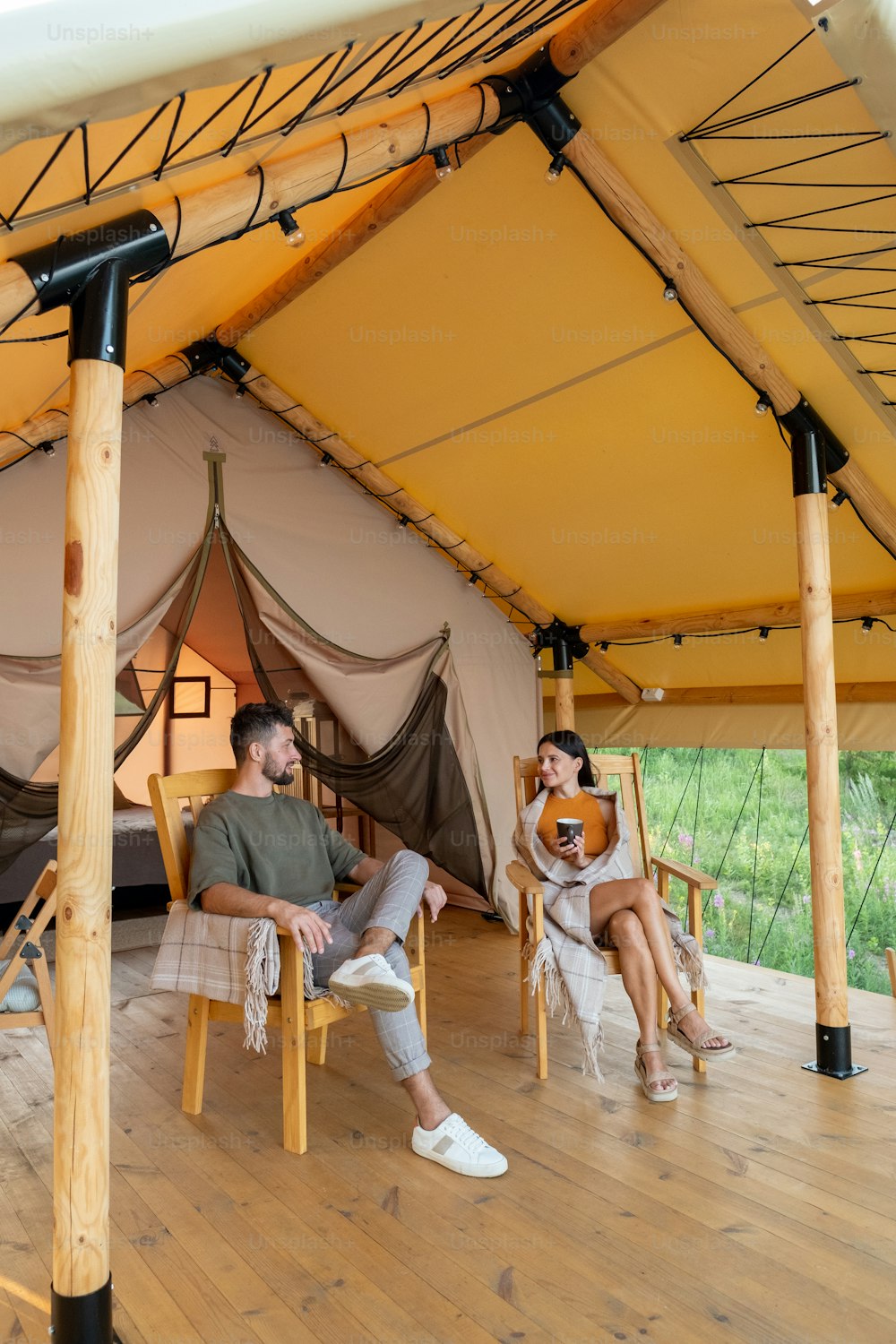 Young restful man and woman talking while relaxing in armchairs by glamping house