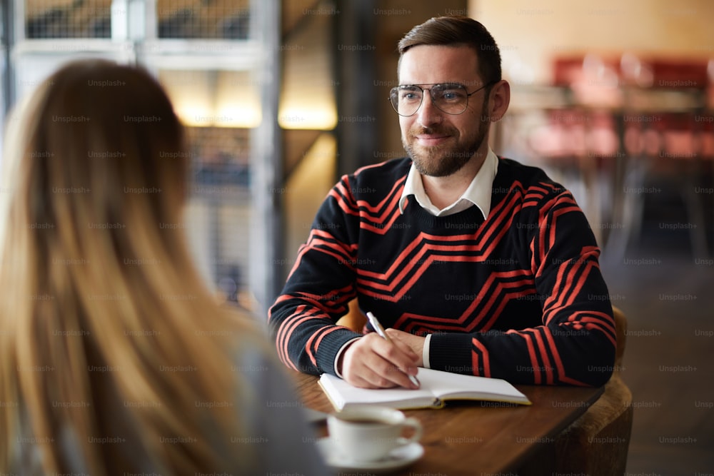 Young confident businessman in eyeglasses looking at colleague at meeting while making working notes
