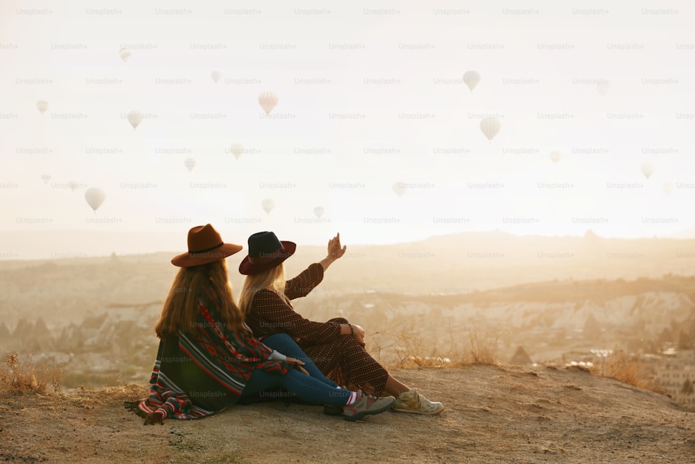 Viajar. Mujeres viajeras que miran globos aerostáticos voladores en el cielo, mujeres con sombreros sentadas en la colina disfrutando de la puesta de sol. Alta resolución