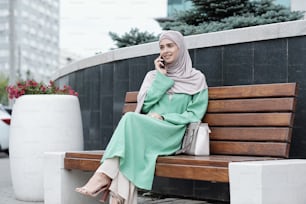 Giovane donna d'affari musulmana elegante sorridente in abito verde lungo e hijab seduta sulla panchina con borsa e chiamata per telefono