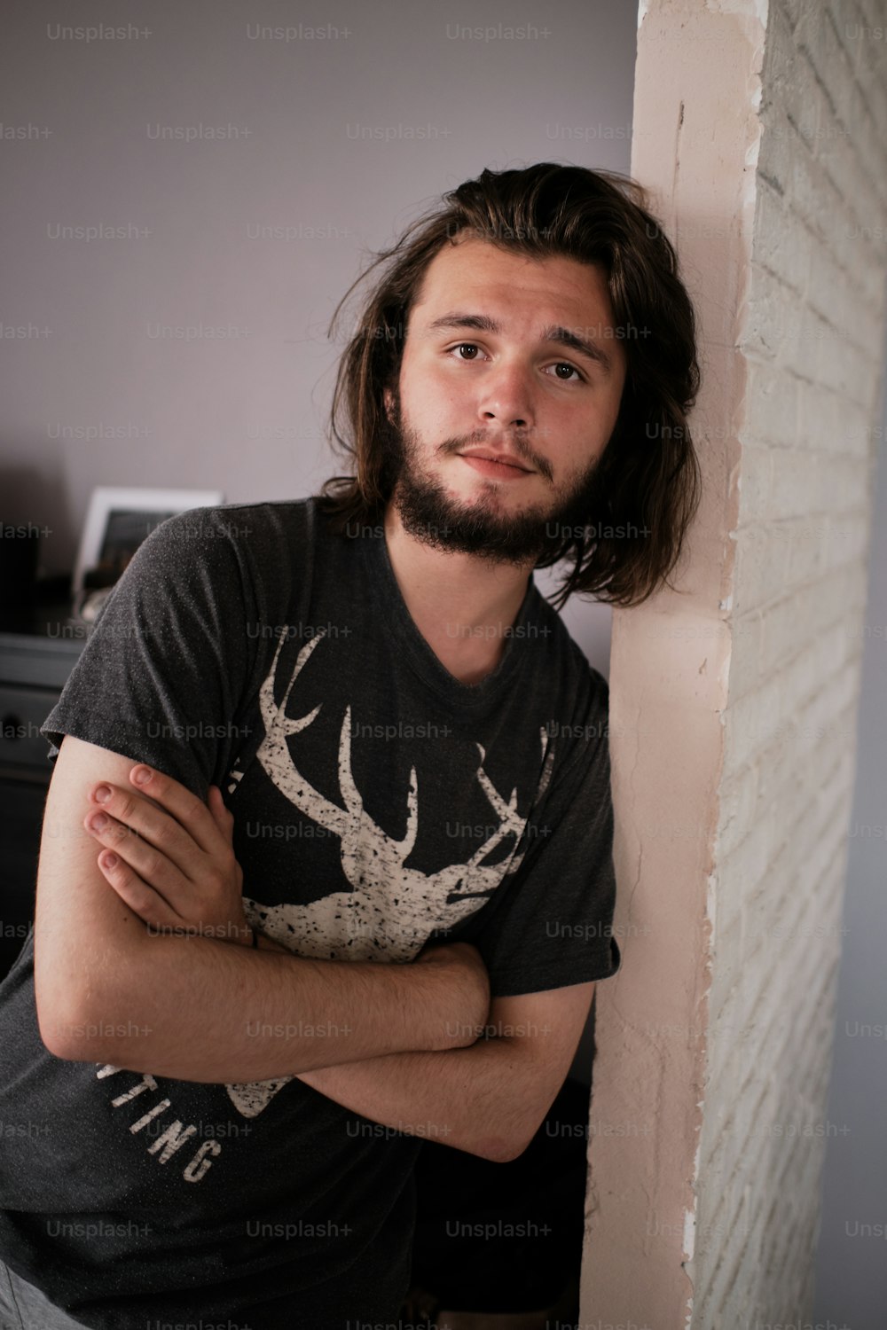 Portrait of young man with beard and long hair. He is standing at the wall, leaning, hands crossed on the chest