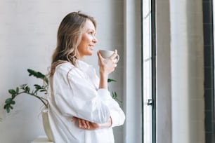 Young smiling blonde woman with long hair in white shirt rest and drinking tea near window in bright modern office