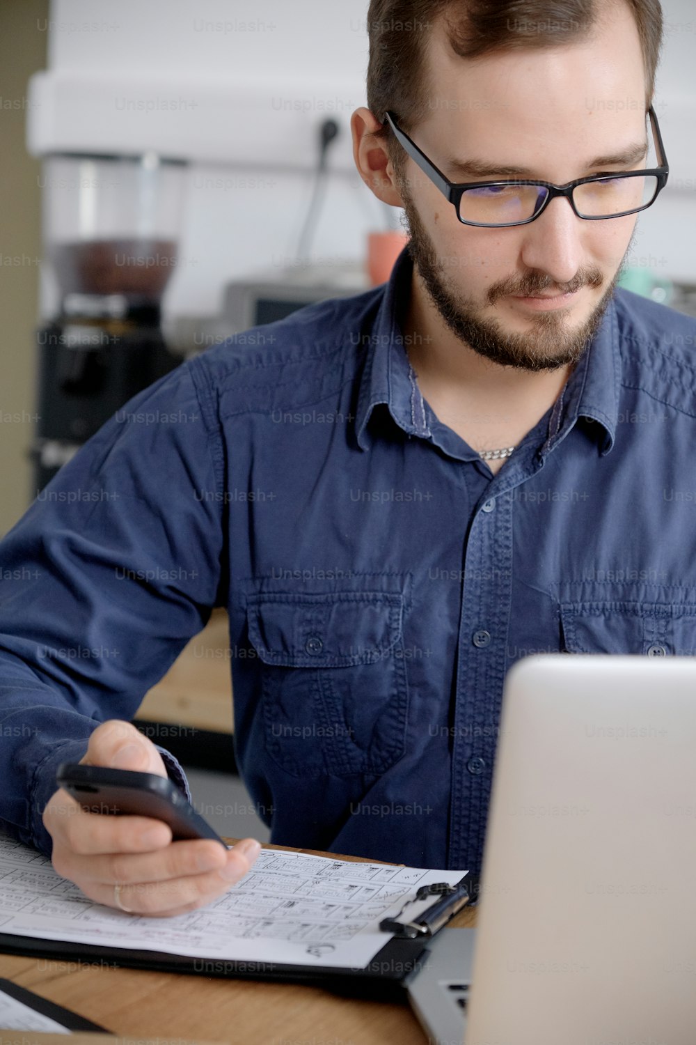 Ritratto di uomo serio in occhiali e camicia blu che lavora su laptop e smartphone mentre si siede al tavolo di legno contro la parete bianca