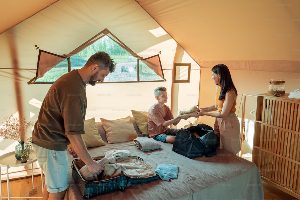 Teenage boy helping his parents with unpacking baggage in bedroom of glamping house