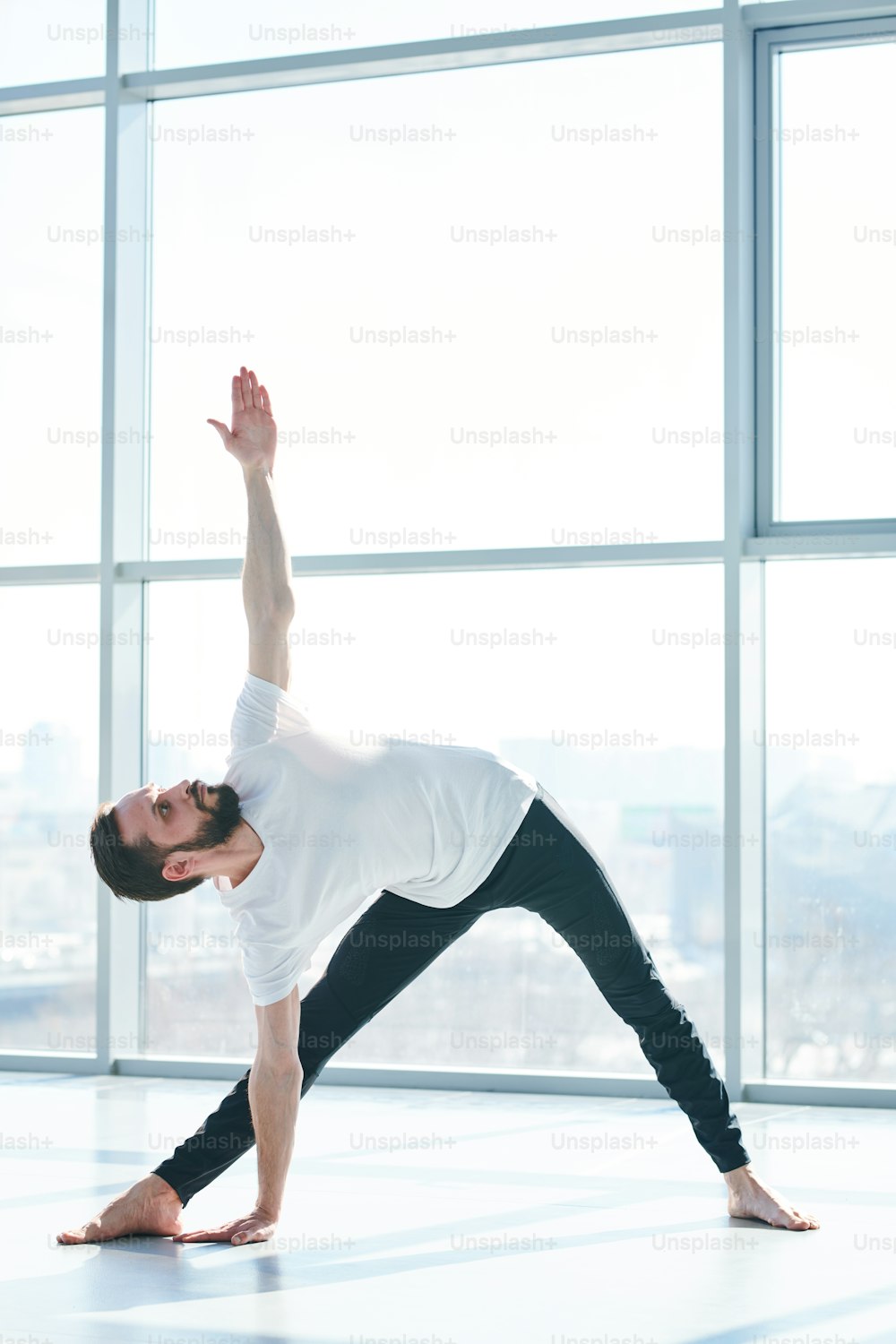 Joven hombre activo con piernas extendidas que se doblan lateralmente con el brazo izquierdo levantado mientras hace ejercicio en el gimnasio