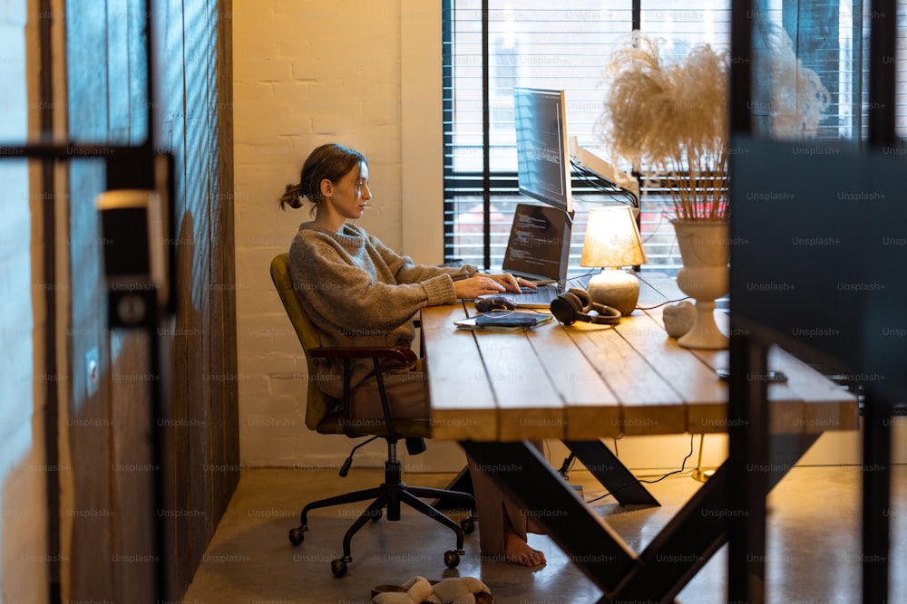 Young woman works on computers, sitting at workplace at cozy home office interior. Concept of freelance and remote work from home. Programmer writing code. Caucasian woman wearing domestic clothes.