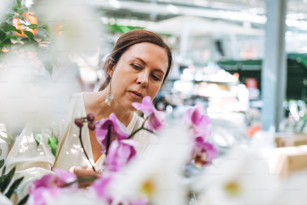 Brunette middle aged woman in white dress buys lilac orchids potted house plants at garden store