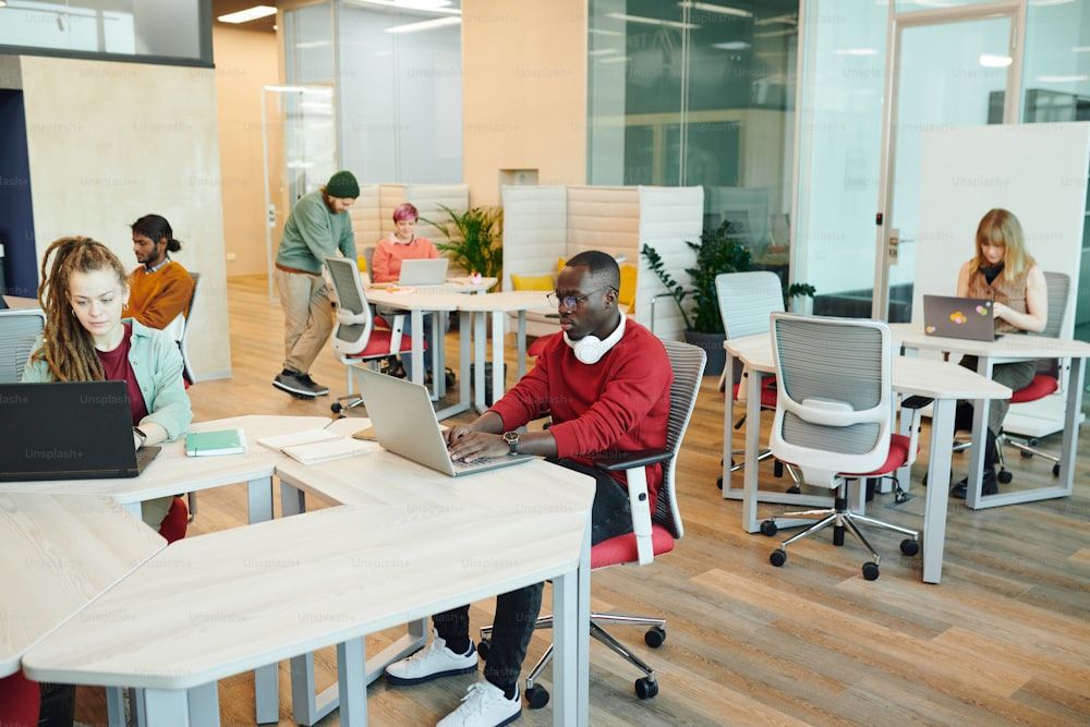 Young serious office managers or designers of various ethnicities sitting by desks and using laptops during individual work in open space