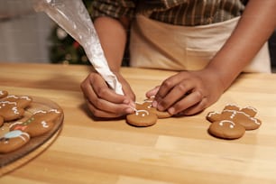 Mains d’une fille africaine décorant des biscuits en pain d’épice faits maison avec un glaçage blanc tout en se tenant près d’une table en bois