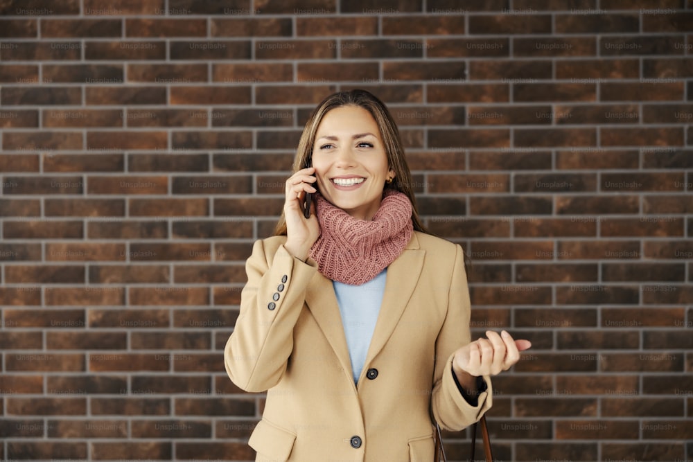 A fashionable businesswoman standing in front of a brick wall and talking on the phone. Business and telecommunications. A businesswoman using the phone.