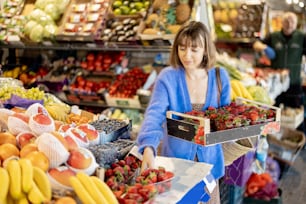 Young woman buying fresh strawberry at local market, standing near the counter full of fruits and berries. Concept of organic local food