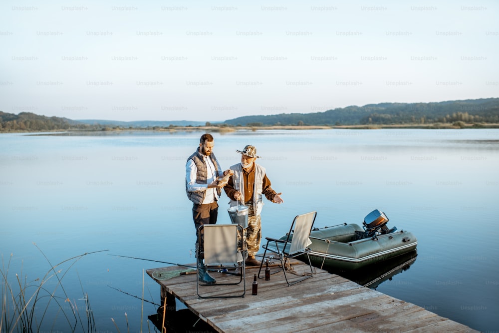 Grandfather with adult son standing together on the wooden pier, enjoying the sunrise while fishing on the lake early in the morning