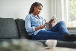 Smiling young African woman using a digital tablet while relaxing alone on her living room sofa at home