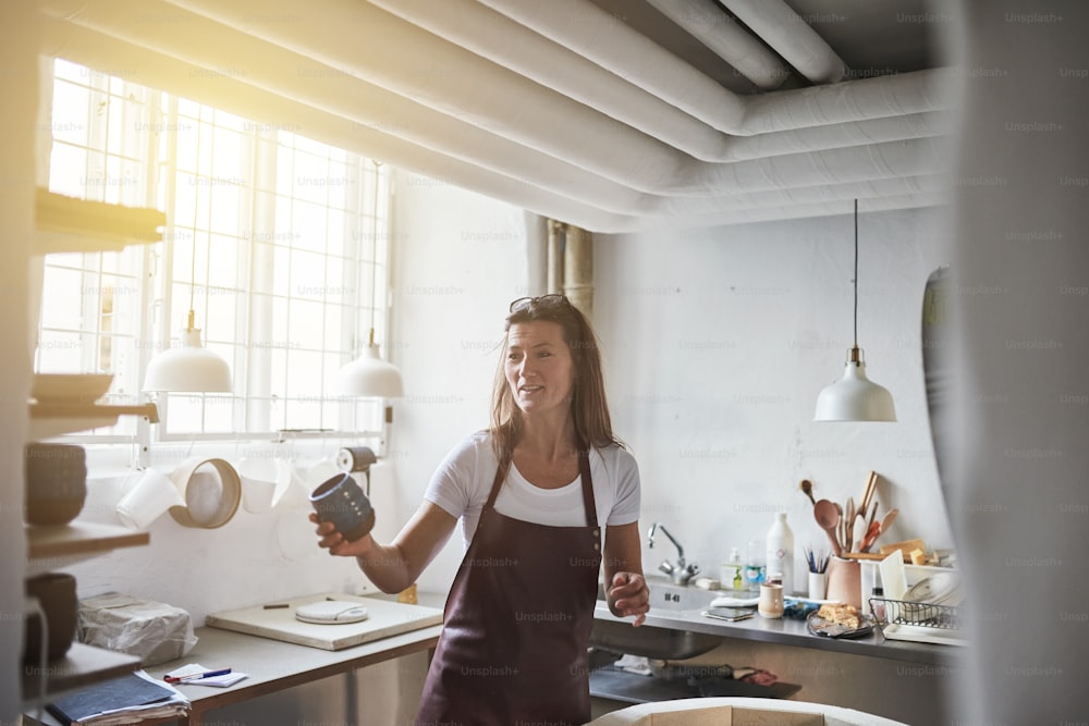 Smiling female artisan standing alone in her creative workshop holding a newly made piece of glazed pottery