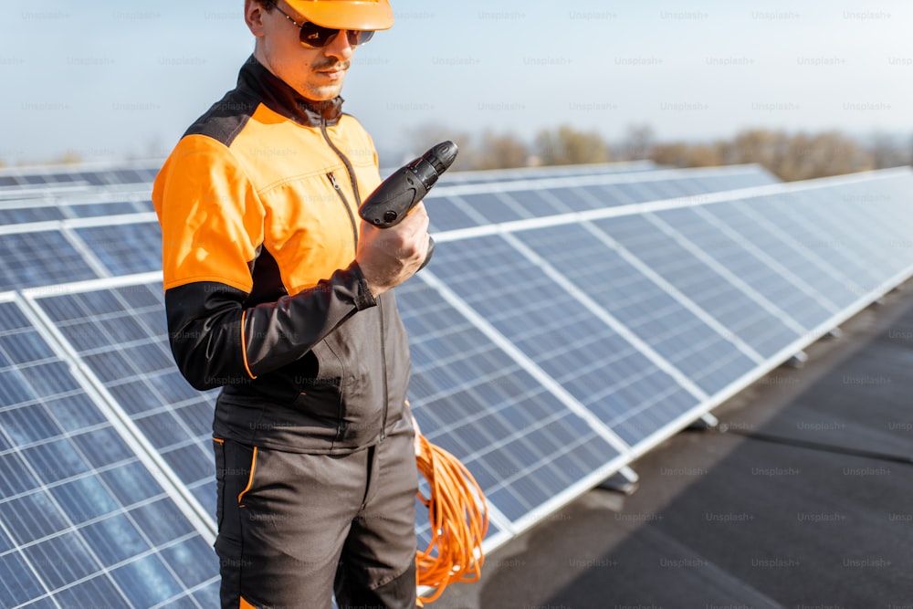 Gut ausgerüsteter Arbeiter in orangefarbener Schutzkleidung bei der Wartung von Sonnenkollektoren auf einer Photovoltaik-Dachanlage. Konzept der Wartung und Installation von Solarstationen